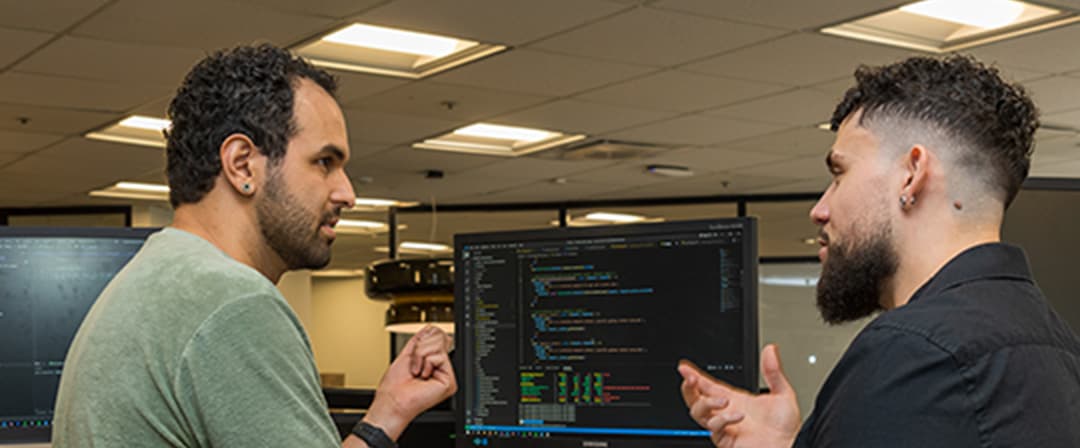 Two software developers discuss code on a computer monitor in an office, gesturing toward the screen with focused, collaborative expressions.