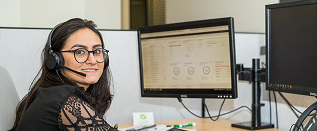 Smiling woman wearing glasses and a headset works at a computer workstation with dual monitors in an office, conveying a friendly, focused mood.