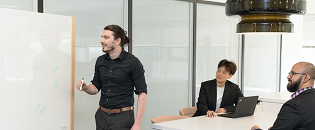 Business team in a modern office: a man presents beside a whiteboard while two colleagues listen at a table, creating a focused, collaborative mood.