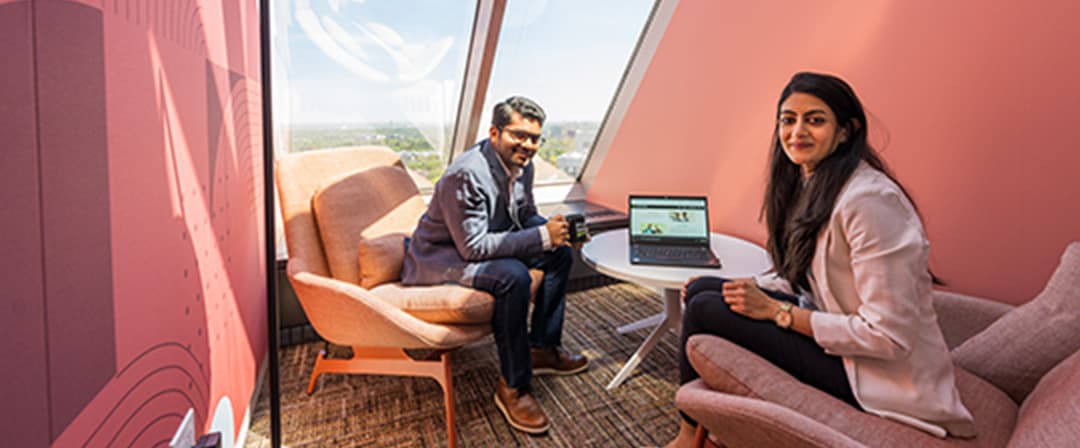 Two coworkers collaborate in a stylish pink office lounge, seated near a window with a laptop, conveying a warm, modern workplace mood.