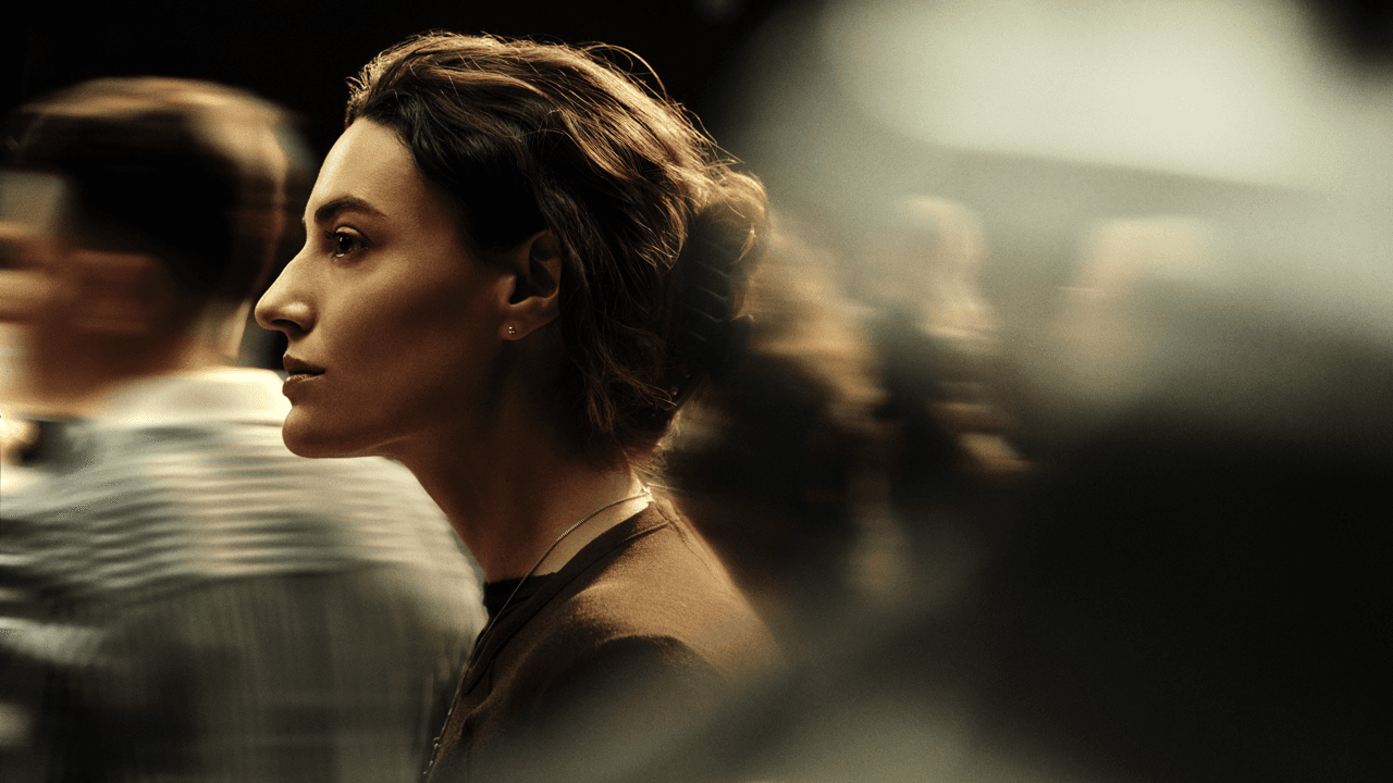 Woman with short wavy hair in profile, calmly looking ahead amid a blurred crowd, lit warmly against a dark background.