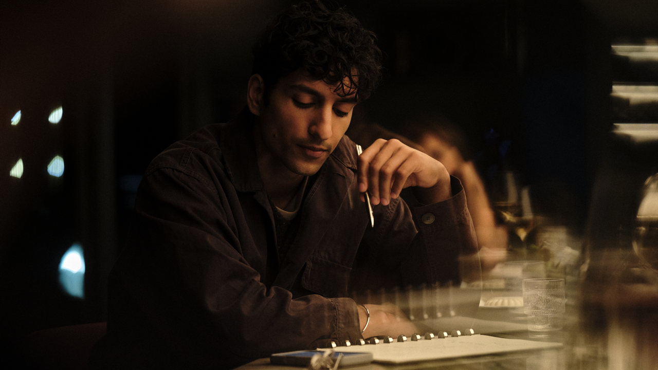 Man with curly hair sitting at a bar or table, holding a pen to his chin while looking down thoughtfully at a spiral notebook, with a phone and glass nearby in warm, dim lighting.