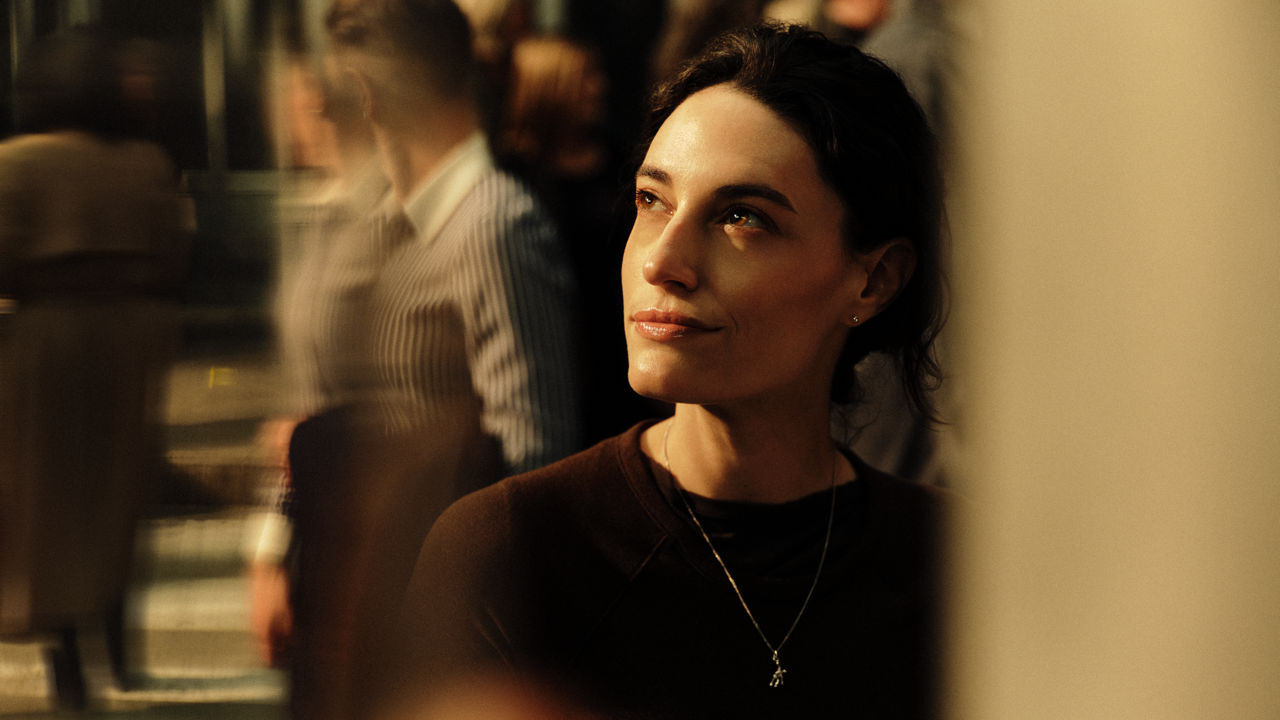 Woman with dark hair pulled back, smiling faintly and looking upward, warmly lit against a blurred crowd of commuters in the background.