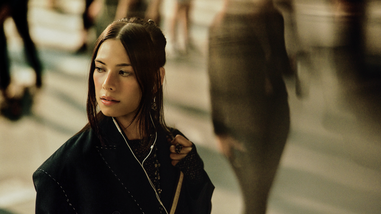 Woman with dark hair and drop earrings wearing earphones and a black coat, glancing to the side in a busy indoor space with blurred passersby in the background.