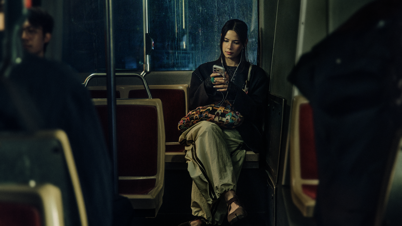 Woman with wet hair sitting alone on a bus at night, wearing earphones and looking at her phone, with a colorful patterned bag on her lap and rain streaking the window behind her.