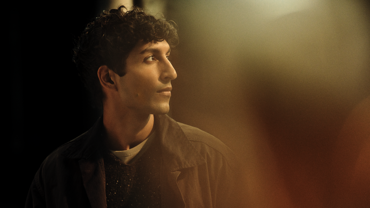 Profile portrait of a young man with curly hair, softly smiling and looking off to the side, warmly lit against a dark, blurred background.