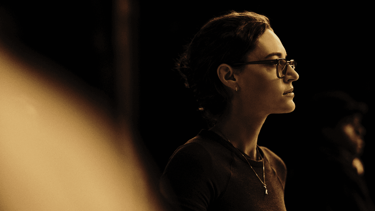 Woman in glasses with short dark hair standing in profile, wearing a necklace, lit warmly against a dark background with another figure blurred behind her.