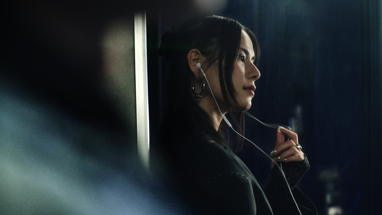 Woman with dark hair and hoop earrings wearing earphones, standing in profile and smiling faintly while adjusting the earphone cord, in dim, moody lighting.