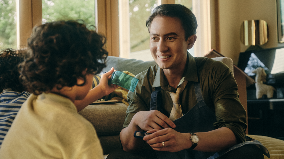 Man in an apron and tie smiling while sitting with a curly-haired child holding up a small phone, in a bright living room with large windows.