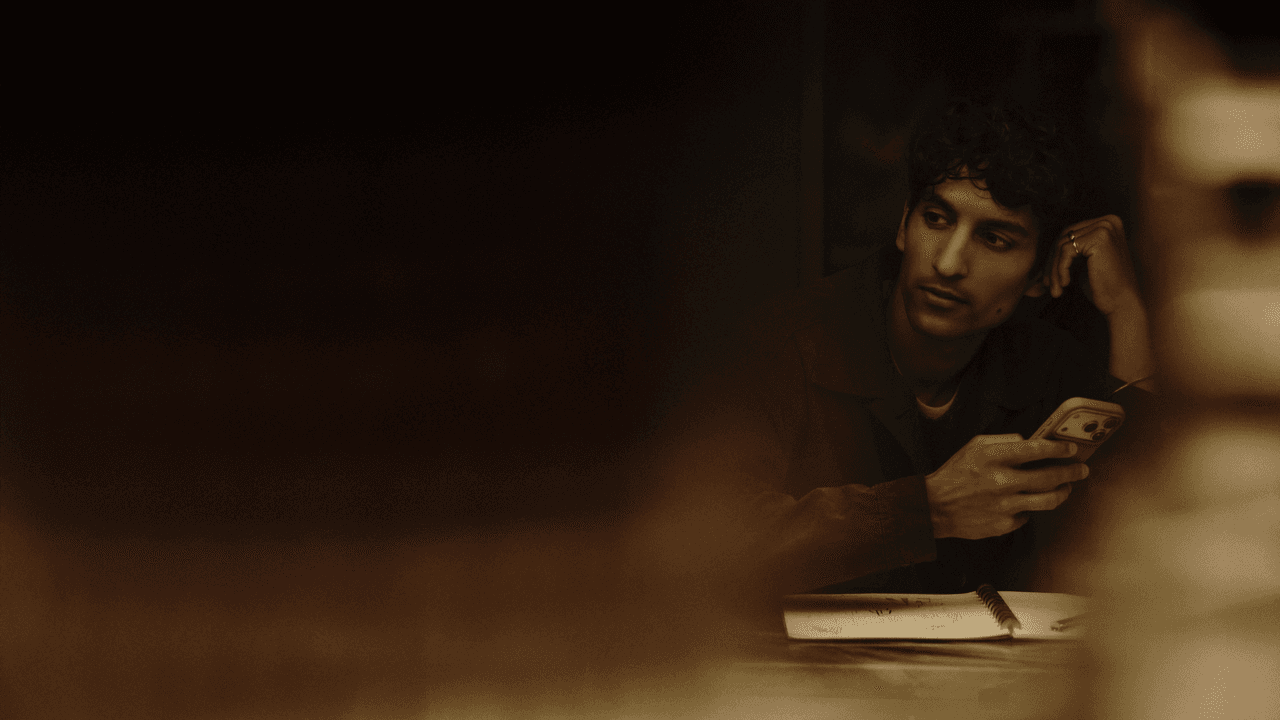 Man with curly hair resting his head on his hand while glancing to the side, holding a phone at a table with an open notebook, in dim, warm lighting.