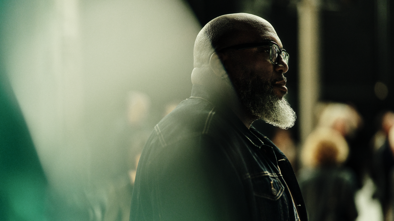 Profile portrait of a bald man with a gray beard and glasses, standing in a denim jacket amid a hazy, softly lit crowd scene.