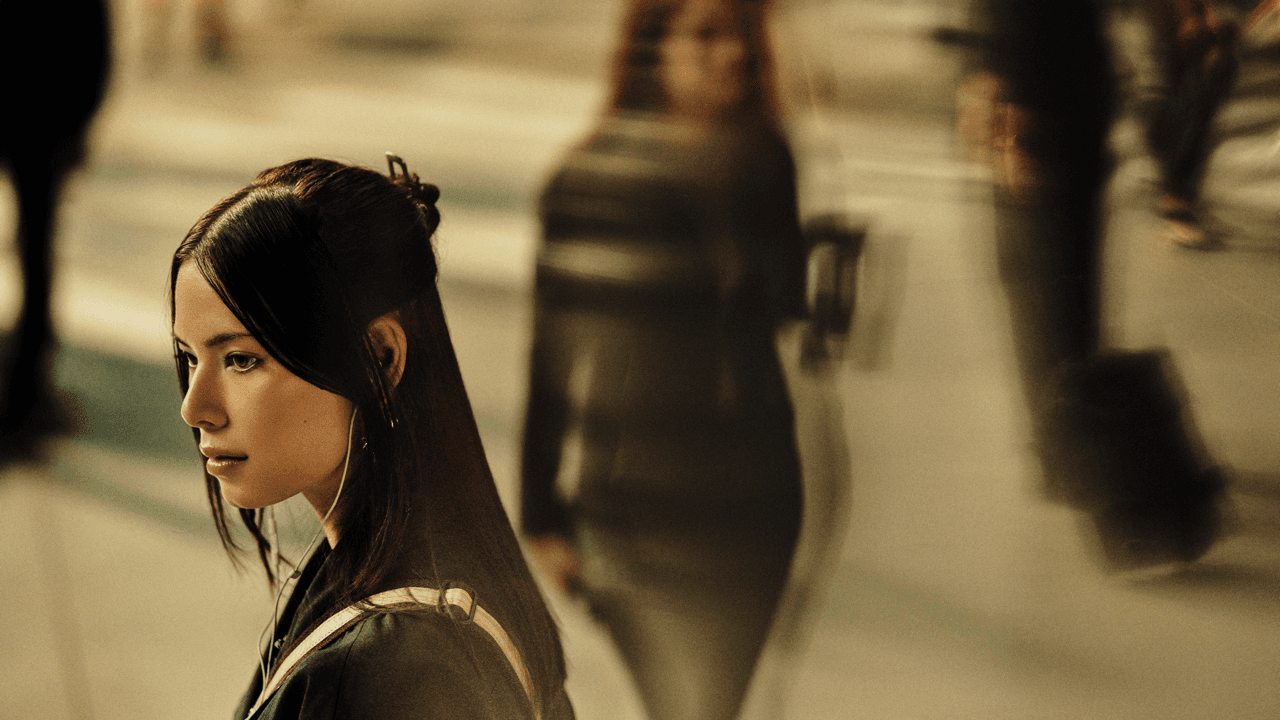 Woman in profile with long dark hair and earphones, standing still amid a blurred crowd of pedestrians on a sunlit street.
