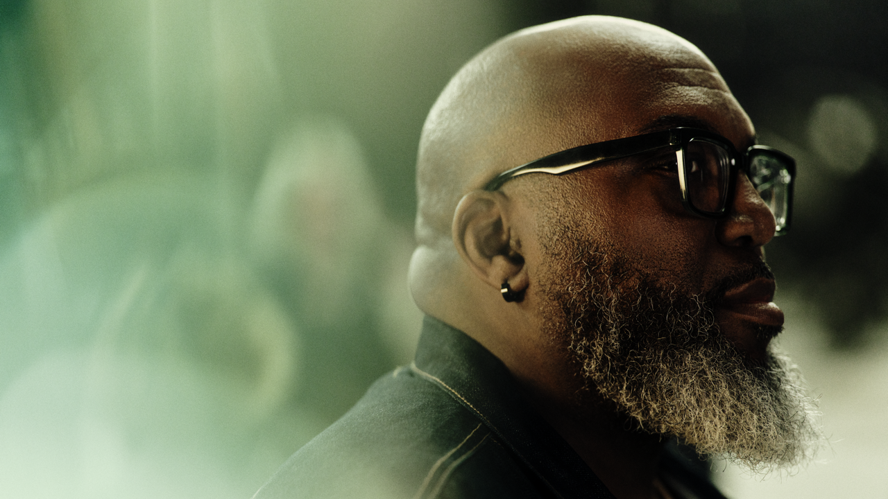 Profile portrait of a bald man with a gray beard and black-framed glasses, wearing an earring, looking off to the side against a soft blurred green background.