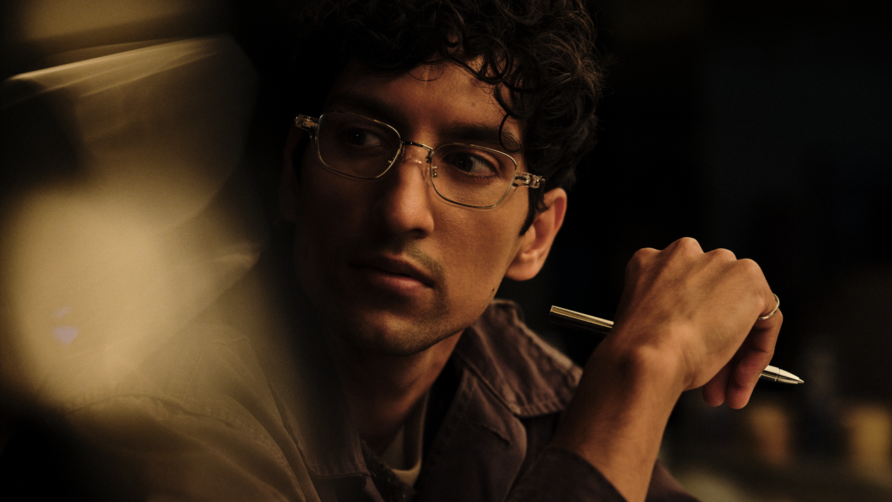 Close-up of a man with curly hair and glasses holding a pen near his shoulder, glancing sideways with a pensive expression in warm, dim lighting.