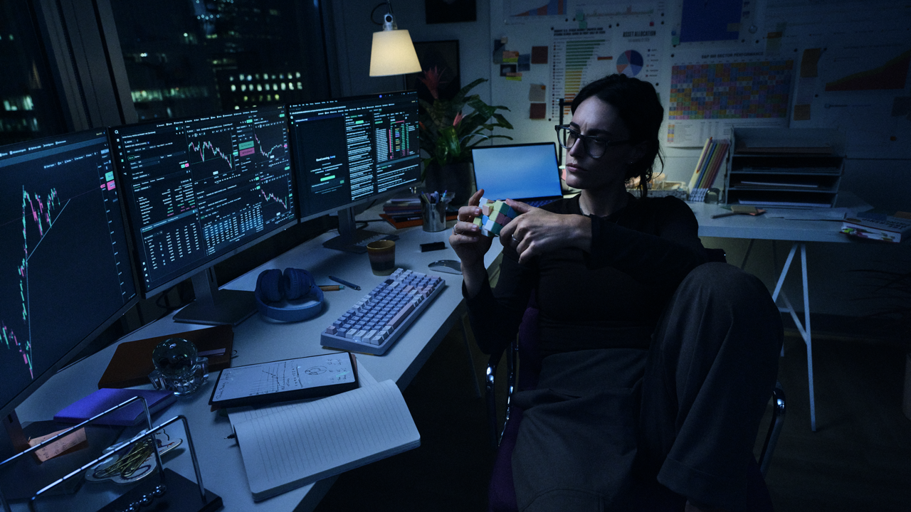 Woman in glasses sitting back in her chair holding a Rubik's cube, taking a break at a desk with multiple monitors showing stock charts, in a dimly lit office at night.