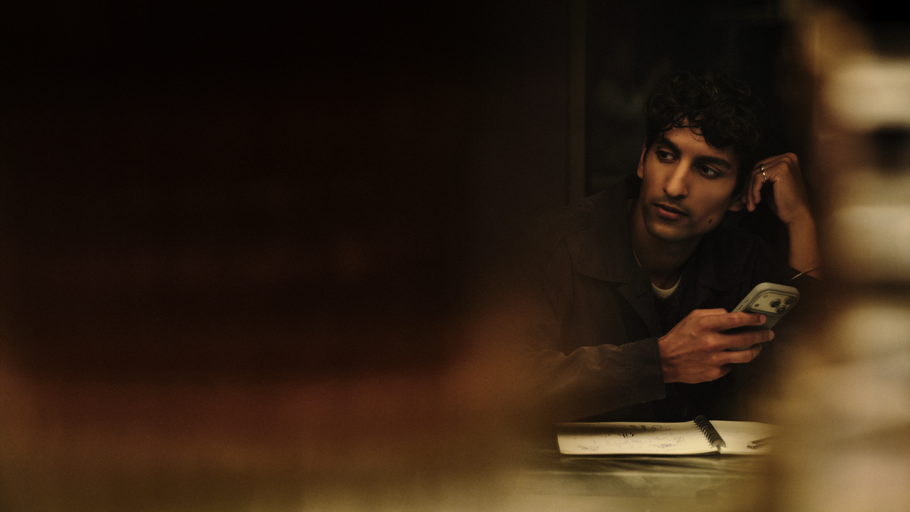 Man with curly hair resting his head on his hand while glancing to the side, holding a phone at a table with an open notebook, in dim, warm lighting.