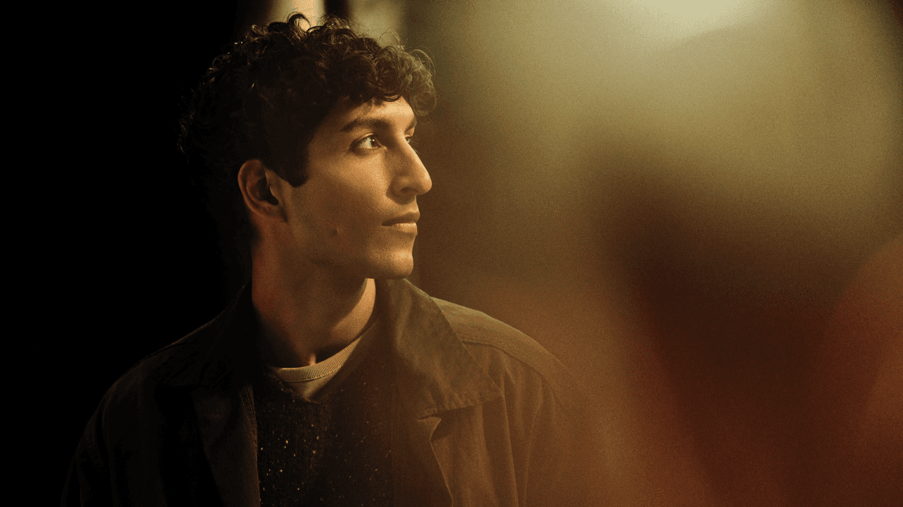 Profile portrait of a young man with curly hair, softly smiling and looking off to the side, warmly lit against a dark, blurred background.