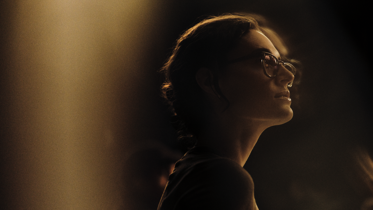 Woman in glasses seen in profile, smiling faintly with her head tilted up, warmly lit against a dark background.