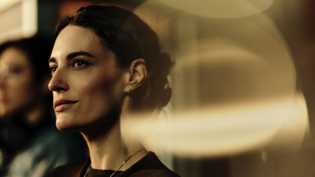Close-up profile of a woman with dark hair pulled back, softly smiling and looking off into the distance, backlit by warm golden light with another person blurred in the background.