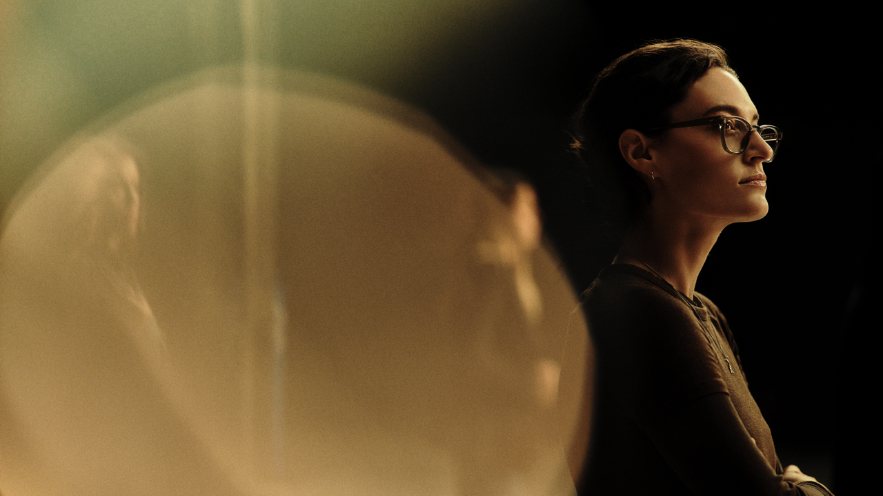 Woman with short dark hair and glasses in profile, arms crossed, gazing off to the side against a dark background with a warm circular light flare in the foreground.