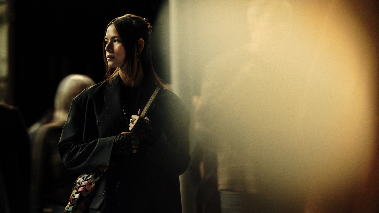 Woman in a dark coat holding the strap of a woven multicolor bag, standing in profile in a dim hallway with a hazy warm light and blurred figures nearby.