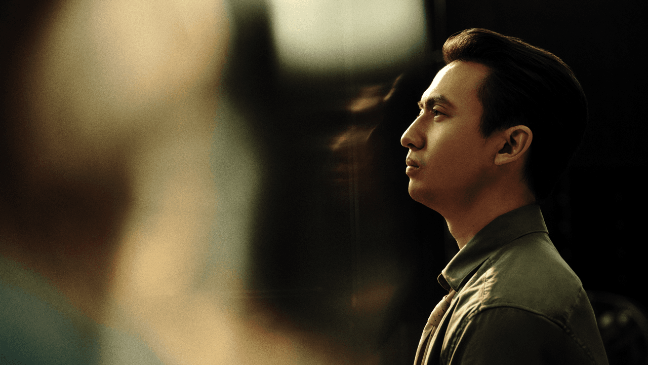 Profile portrait of a man with slicked-back hair wearing a green jacket, gazing forward with a serious expression against a dark, softly reflective background.