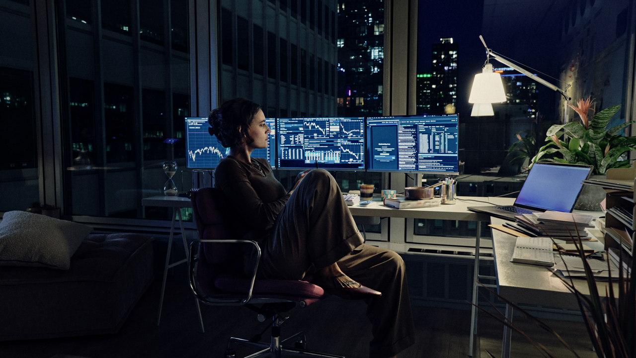 Woman sitting cross-legged in an office chair, viewing stock charts on three monitors in a home office at night, with a city skyline visible through large windows and a desk lamp, laptop, and plants nearby.