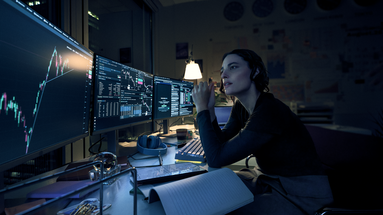 Woman with hands clasped studying multiple monitors showing candlestick charts and trading data at a dimly lit desk, with headphones, a notebook, and a desk lamp nearby at night.