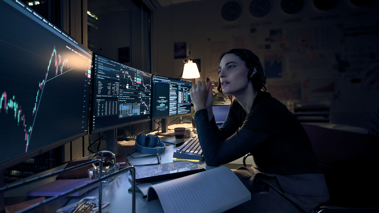 Woman with hands clasped studying multiple monitors showing candlestick charts and trading data at a dimly lit desk, with headphones, a notebook, and a desk lamp nearby at night.