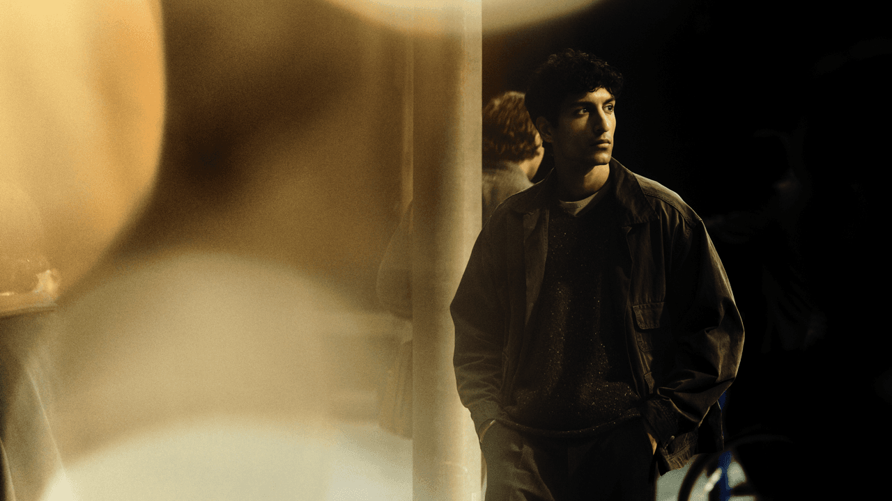 Man with curly hair standing with hands in pockets, looking to the side in a dim, warmly lit space with a hazy light flare and a blurred figure behind him.