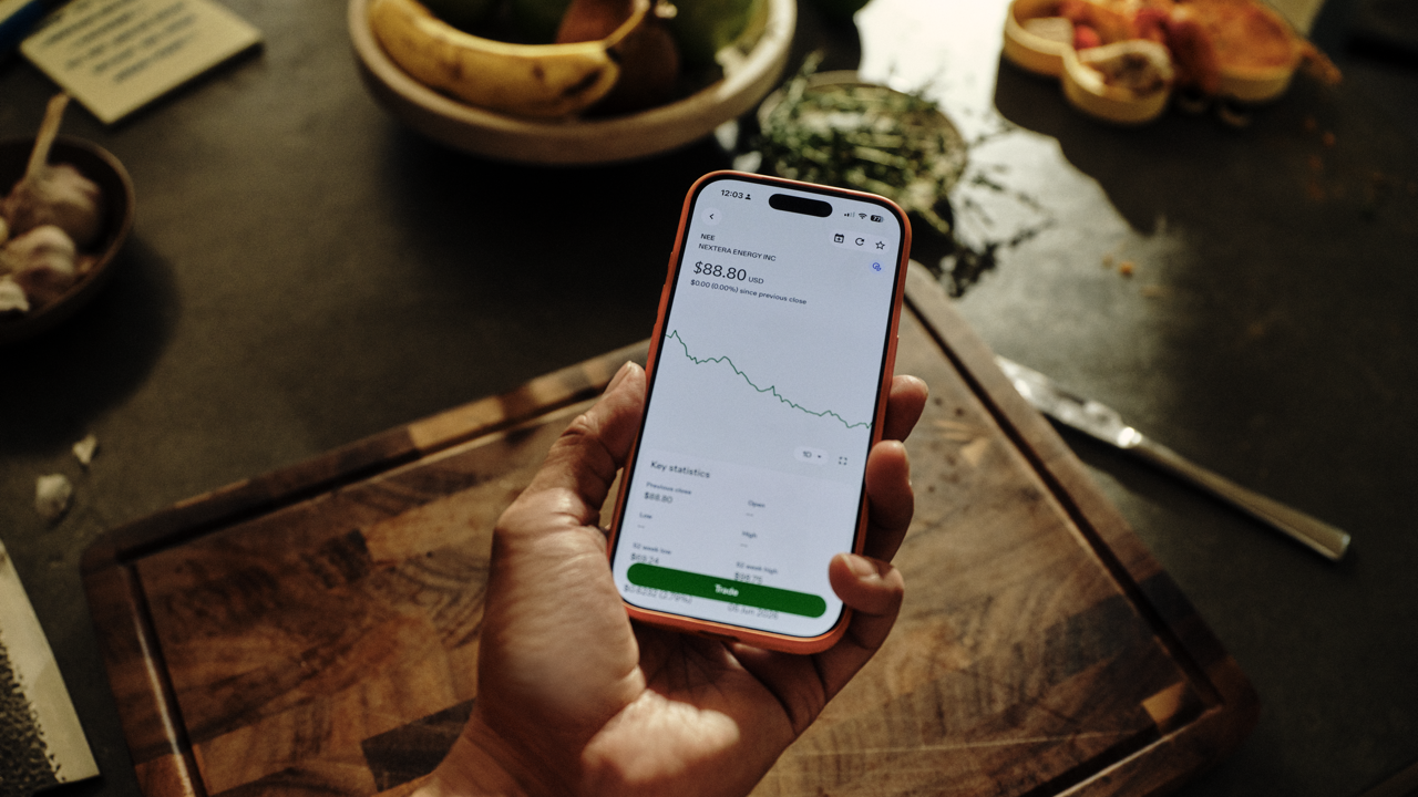 Hand holding a phone displaying a stock quote and price chart for NextEra Energy, held over a wooden cutting board in a kitchen with fruit and food in the background.