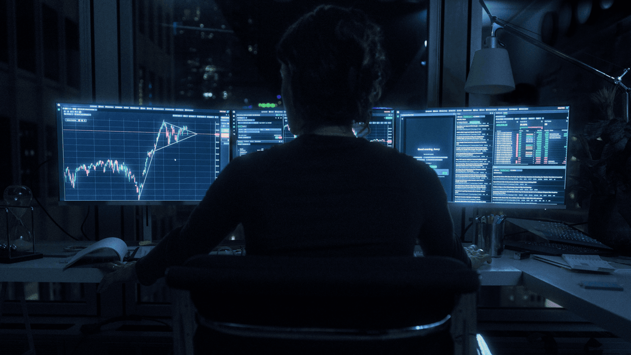 Silhouette of a person seated at a desk, facing three monitors displaying candlestick charts and market data, with a city skyline visible through the window at night.
