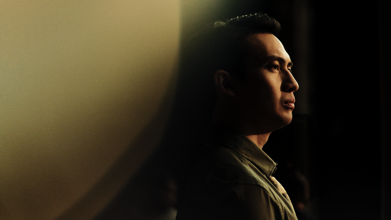 Profile portrait of a man in a green jacket standing in shadow, his face lit by warm side light against a dark background.