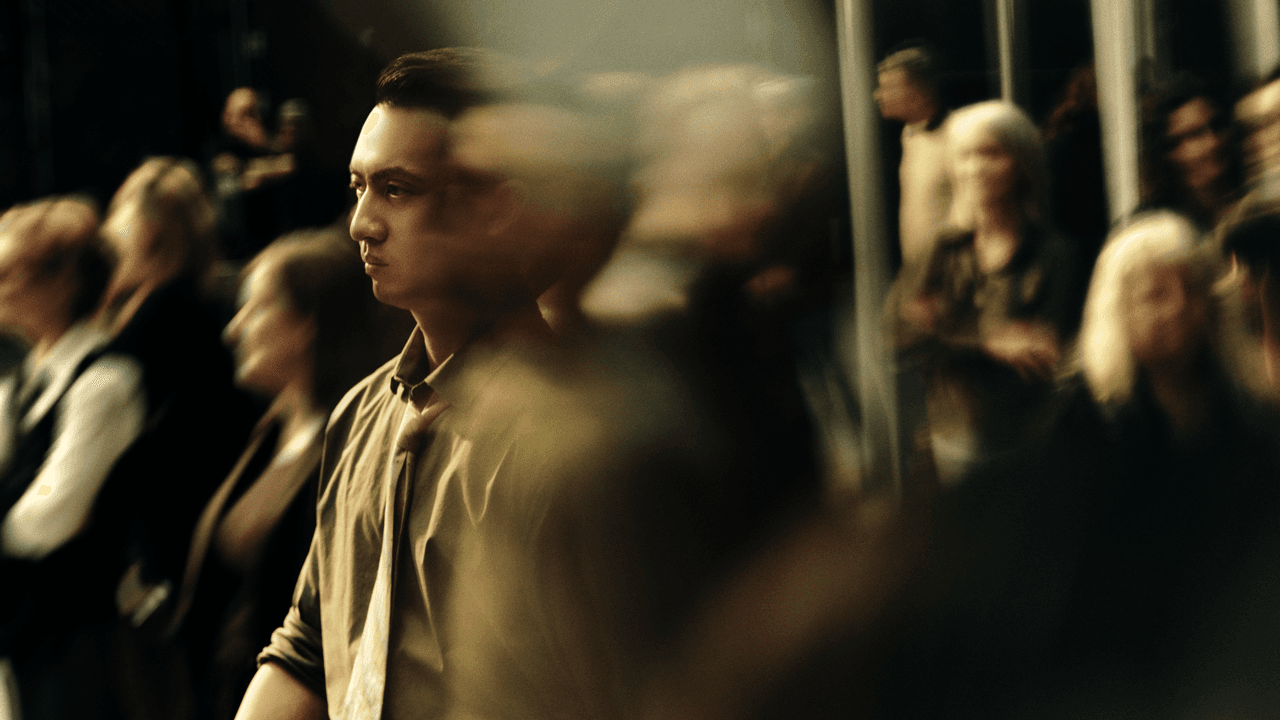 Man in a green shirt and tie in profile, motion-blurred as he moves through a crowd of people seated or standing nearby.