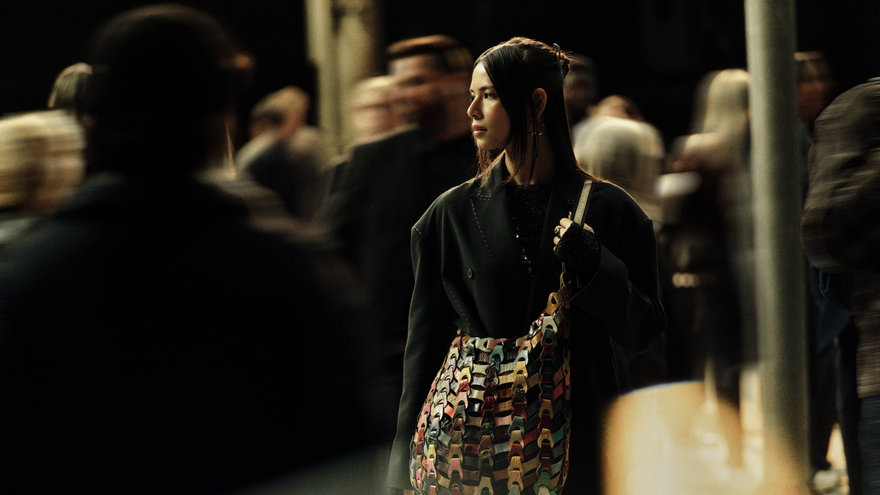 Woman in a dark coat carrying a woven multicolor bag, standing in profile amid a crowd of blurred commuters in a dimly lit space.