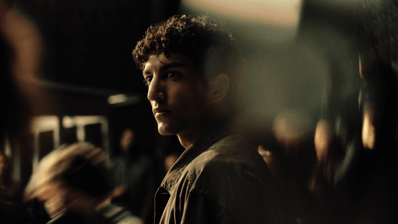 Man with curly hair standing among a crowd, glancing over his shoulder with a slight smile, warmly lit against a dark, blurred background.