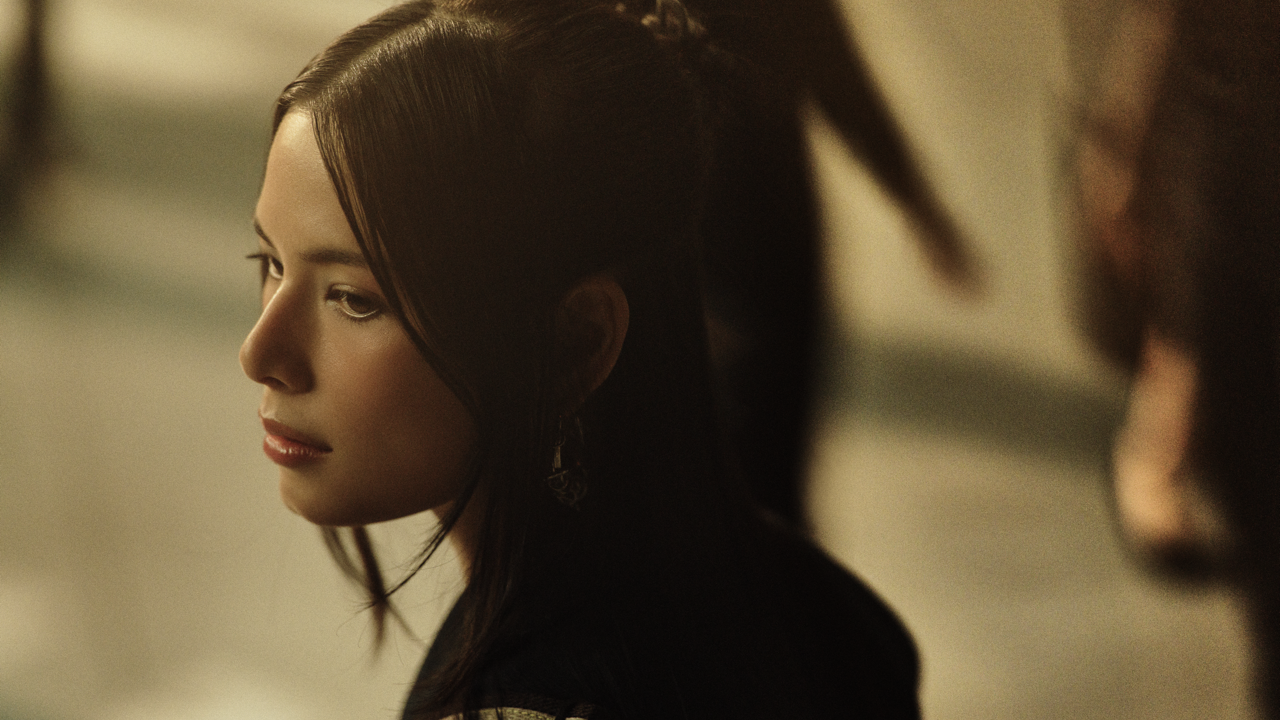 Close-up profile of a woman with long dark hair and drop earrings, looking down thoughtfully, softly lit against a warm, blurred background.