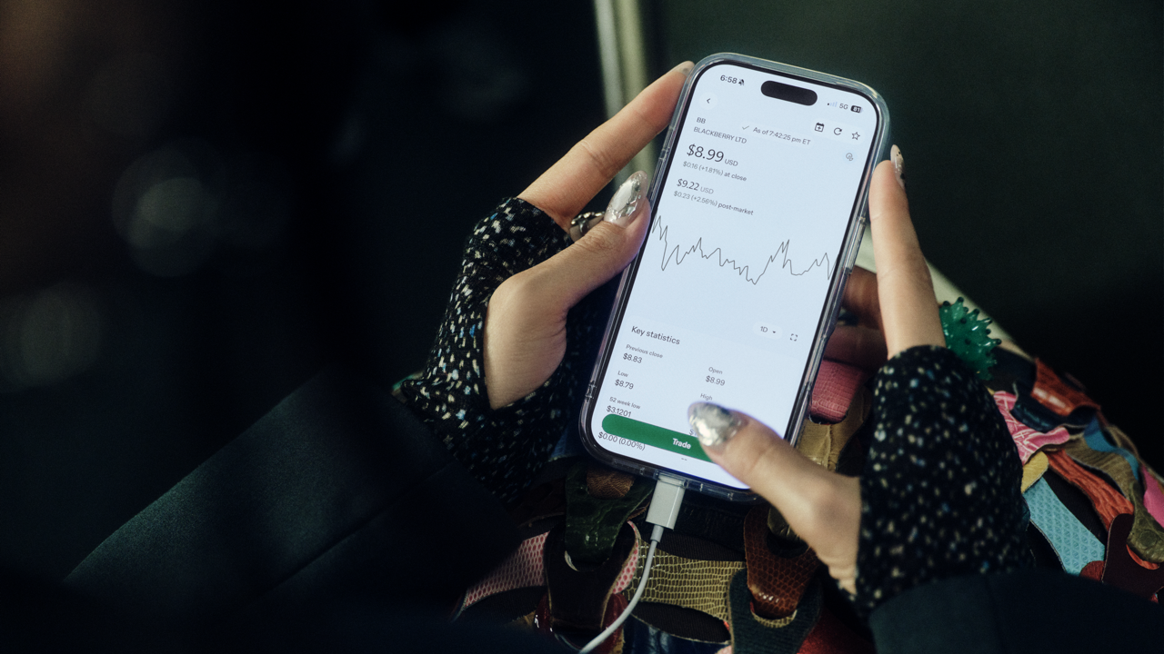 Close-up of hands holding a phone showing BlackBerry Ltd stock quote and price chart, with a patterned bag and dark clothing visible around the phone.