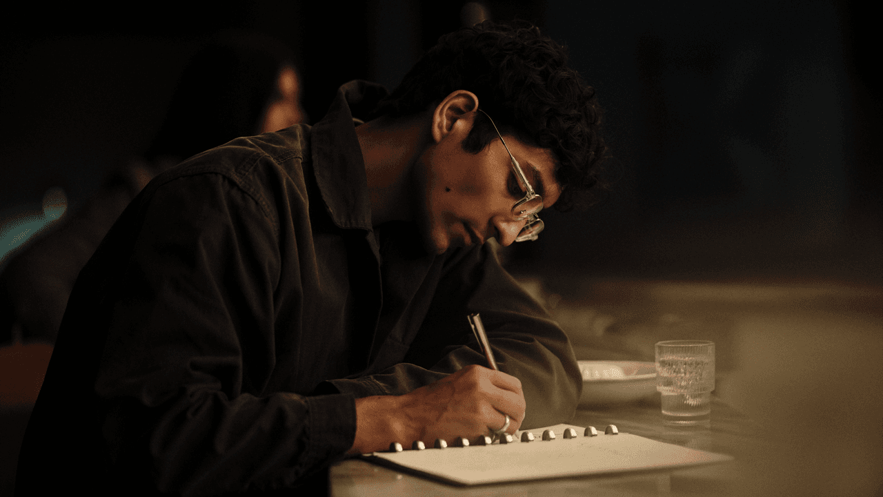 Man with curly hair and glasses leaning over a table, writing intently in a spiral notebook with a pen, a glass of water beside him in warm, dim lighting.