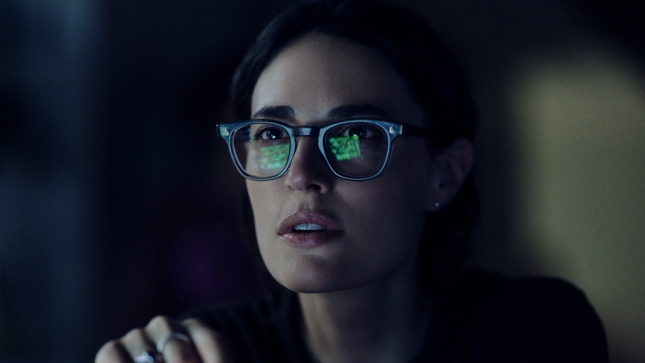 Close-up of a woman with parted lips looking up with a startled expression, green data reflected in her glasses lenses, lit dimly against a dark background.