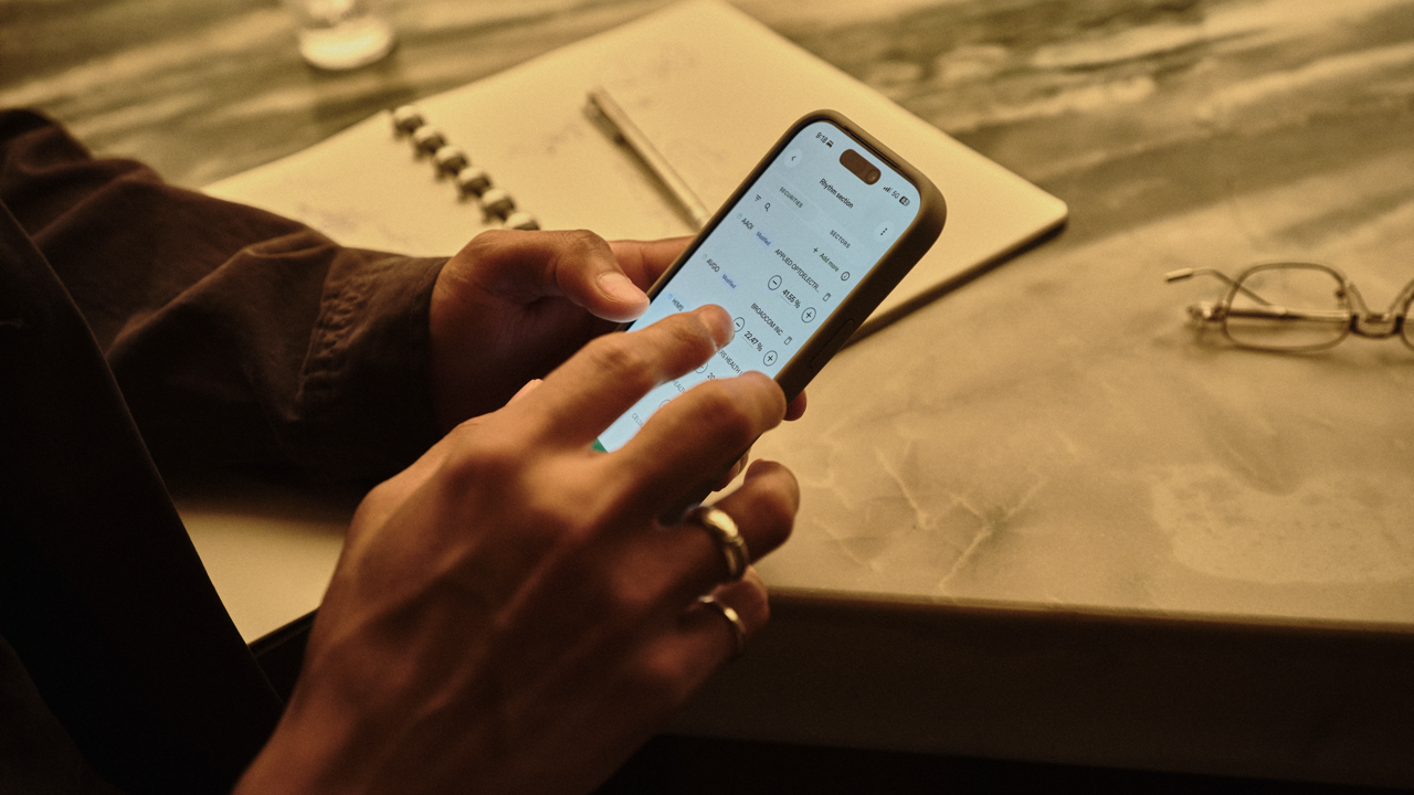 Close-up of hands holding a smartphone displaying a stock portfolio app with holdings and percentage changes, resting on a marble table next to an open notebook, pen, glasses, and a glass of water.