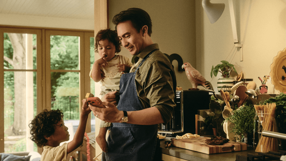 Man in an apron holding a toddler and looking at a phone handed up by a young child in a bright kitchen, with cooking ingredients, herbs, and a decorative bird figurine on the counter.