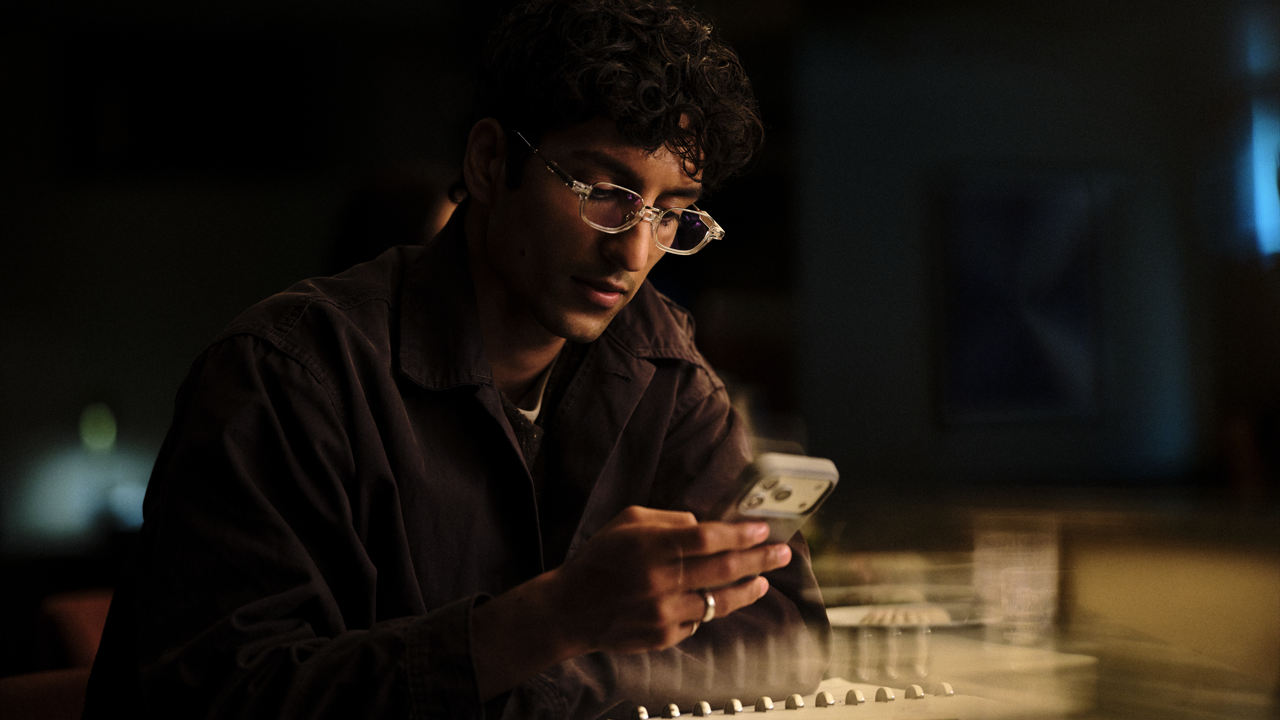 Man with curly hair and clear-framed glasses looking down at his phone, illuminated by its glow while seated at a table in a dimly lit room.