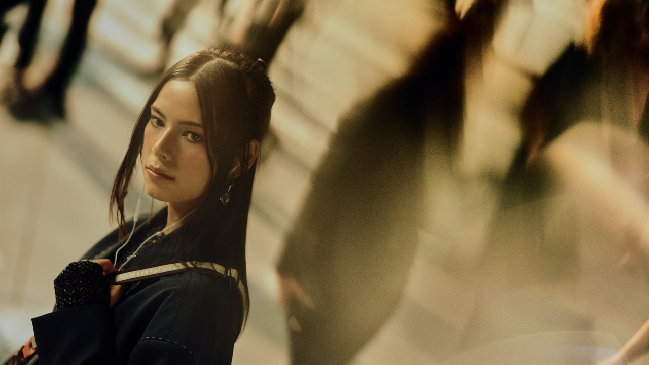 Woman with long dark hair and drop earrings glancing back over her shoulder while wearing earphones, standing in a warmly lit space with blurred figures nearby.