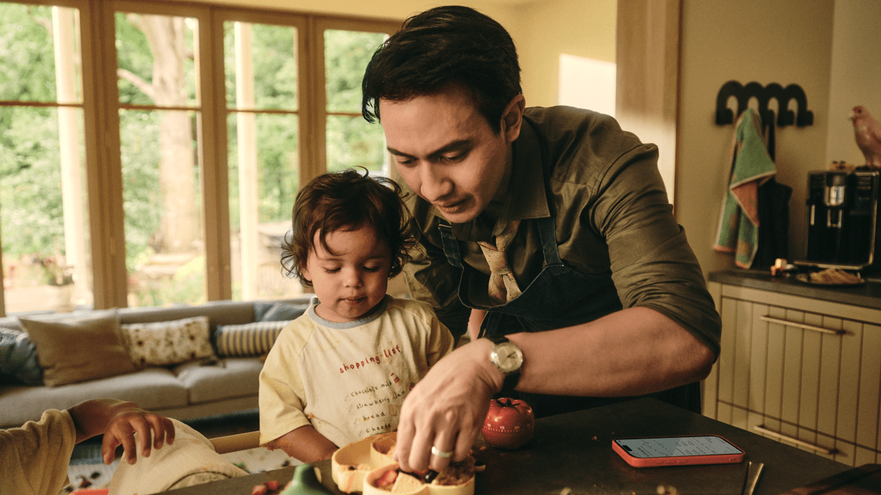 Man in an apron helping a toddler prepare food at a kitchen counter, with a phone and tomato-shaped timer nearby, in a bright home with large windows overlooking greenery.
