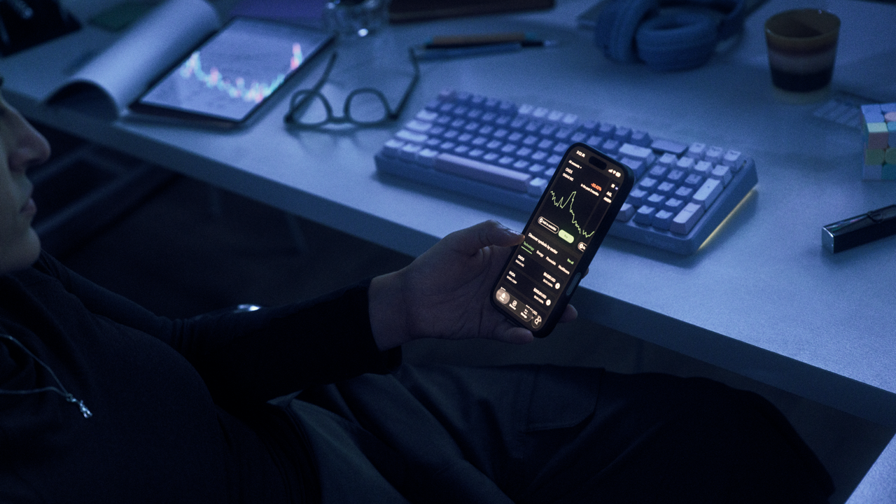 Person holding a phone displaying a stock trading app with charts and market data, at a dimly lit desk with a keyboard, tablet showing a price chart, glasses, and a Rubik's cube.